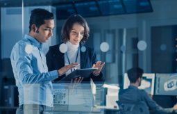 In Technology Research Facility: Female Project Manager Talks With Chief Engineer, they Consult Tablet Computer. Team of Industrial Engineers, Developers Work on Engine Design Using Computers